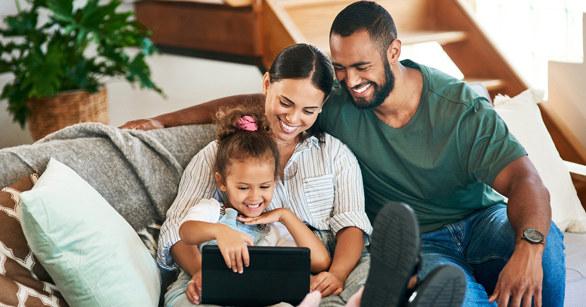 A happy family of three sitting on a sofa, engaged with a tablet.